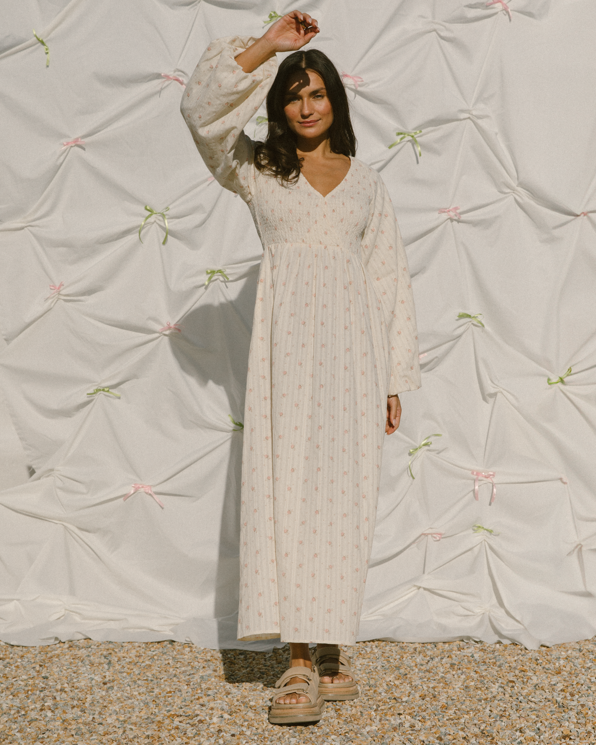 Woman in a long, light-colored dress standing against a textured white wall.