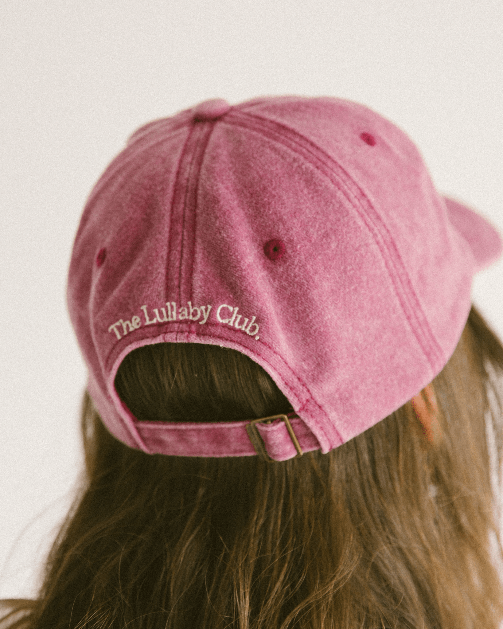 Back of distressed denim cap on womans head in a light maroon colour. With 'The Lullaby Club' embroidered on the back.