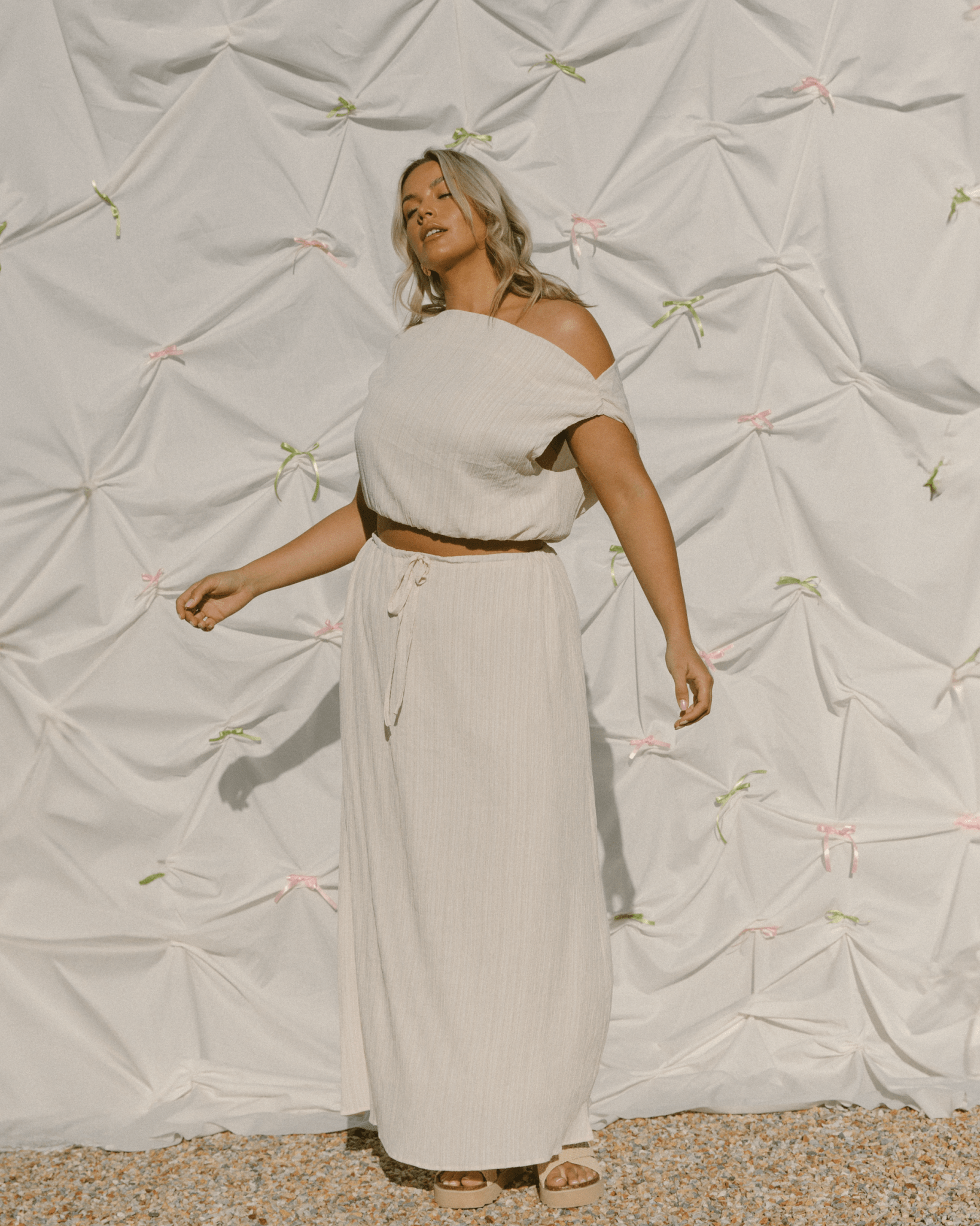 Woman wearing a flowy summer co-ord set in cream colour featuring off the shoulder top and long length skirt.