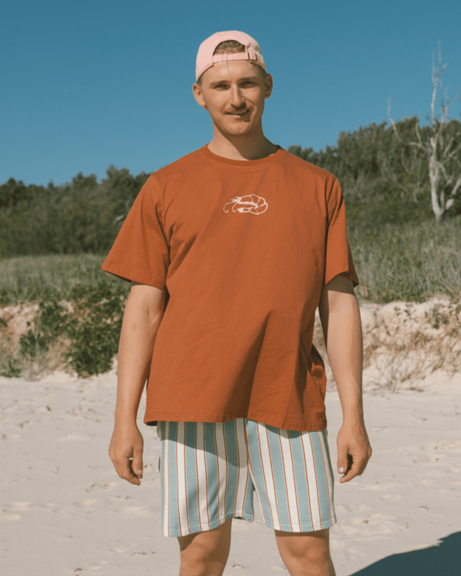 Man wearing an rust t-shirt and striped shorts standing on a sandy beach with trees in the background.