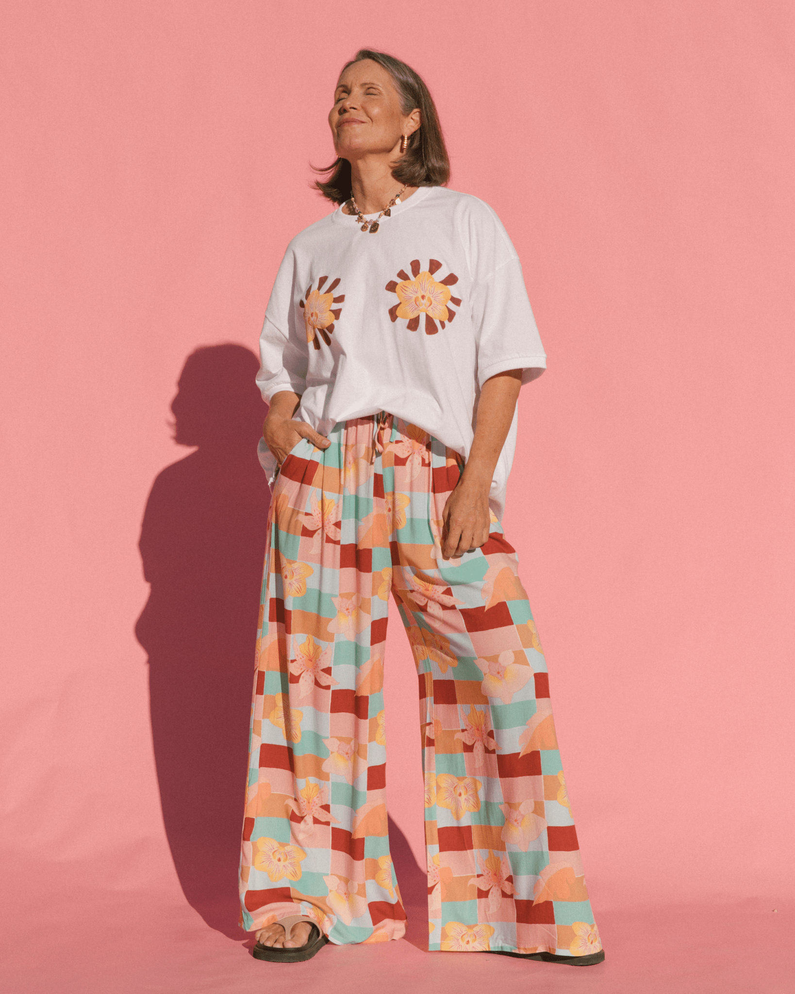 Person wearing a white t-shirt with floral designs and colorful wide-leg pants against a pink background