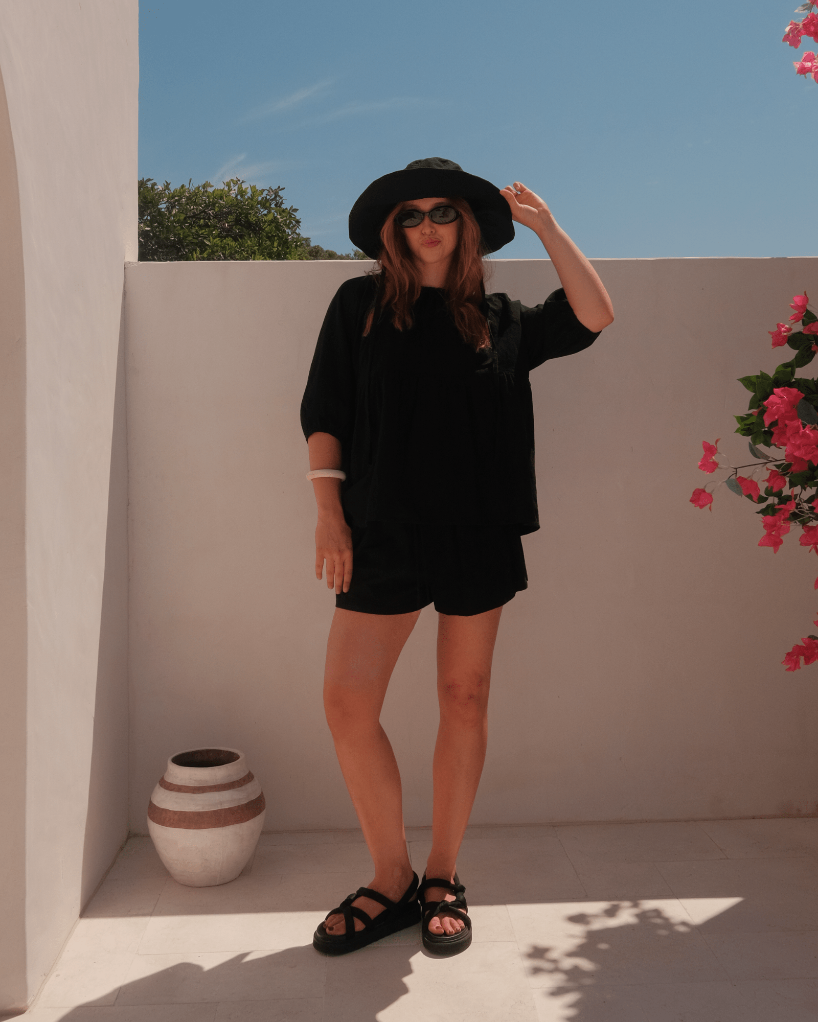 Woman in black outfit with black wide-brimmed hat posing outdoors with a white wall and plant in the background