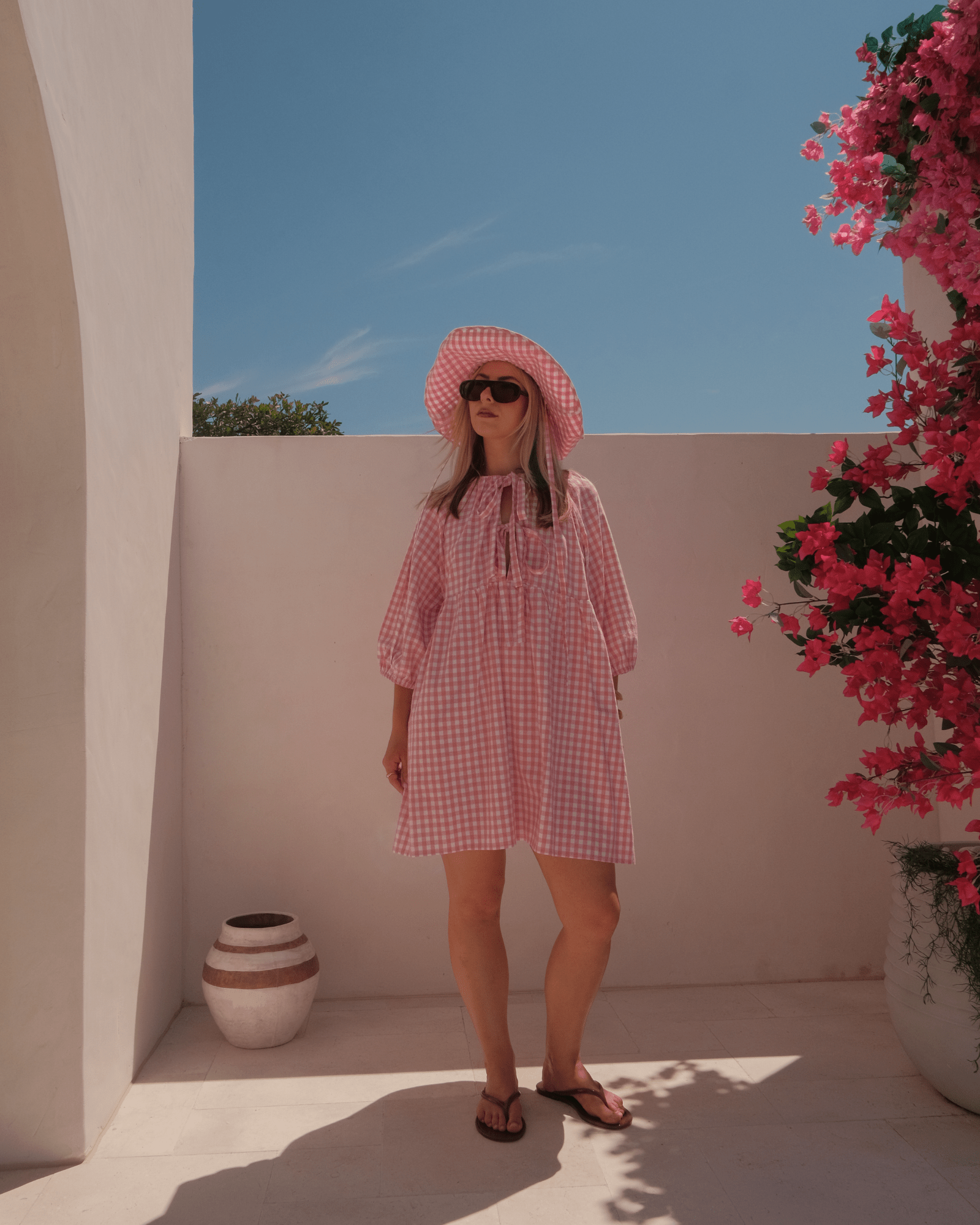 Woman in a pink checkered dress standing outdoors with a clear blue sky and pink flowers.