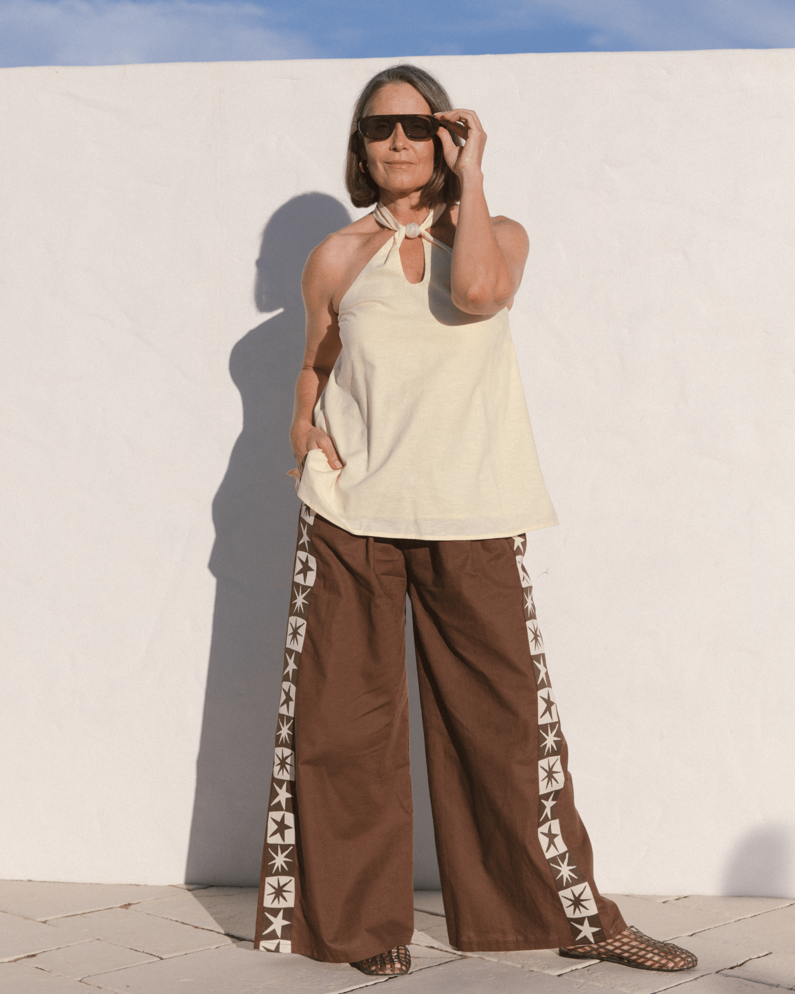 Woman wearing a pale yellow sleeveless top and brown pants with white star patterns against a white wall.