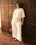 A woman wearing a white short-sleeve top and matching wide-leg pants stands against a dark wood-paneled wall, holding a small red handbag.