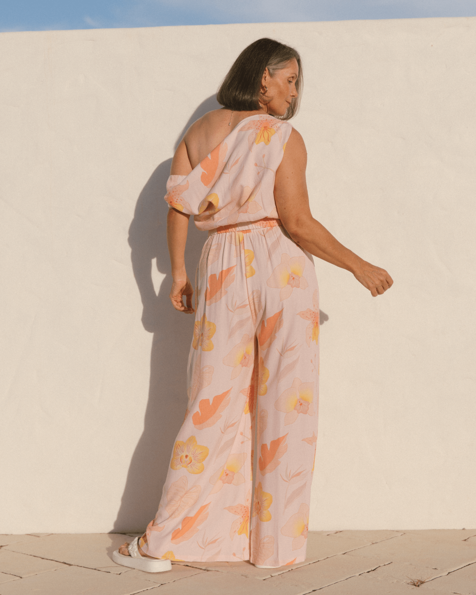Woman showing back of spring outfit, showing off the shoulder crop top & matching long pants in a pink floral pattern. 