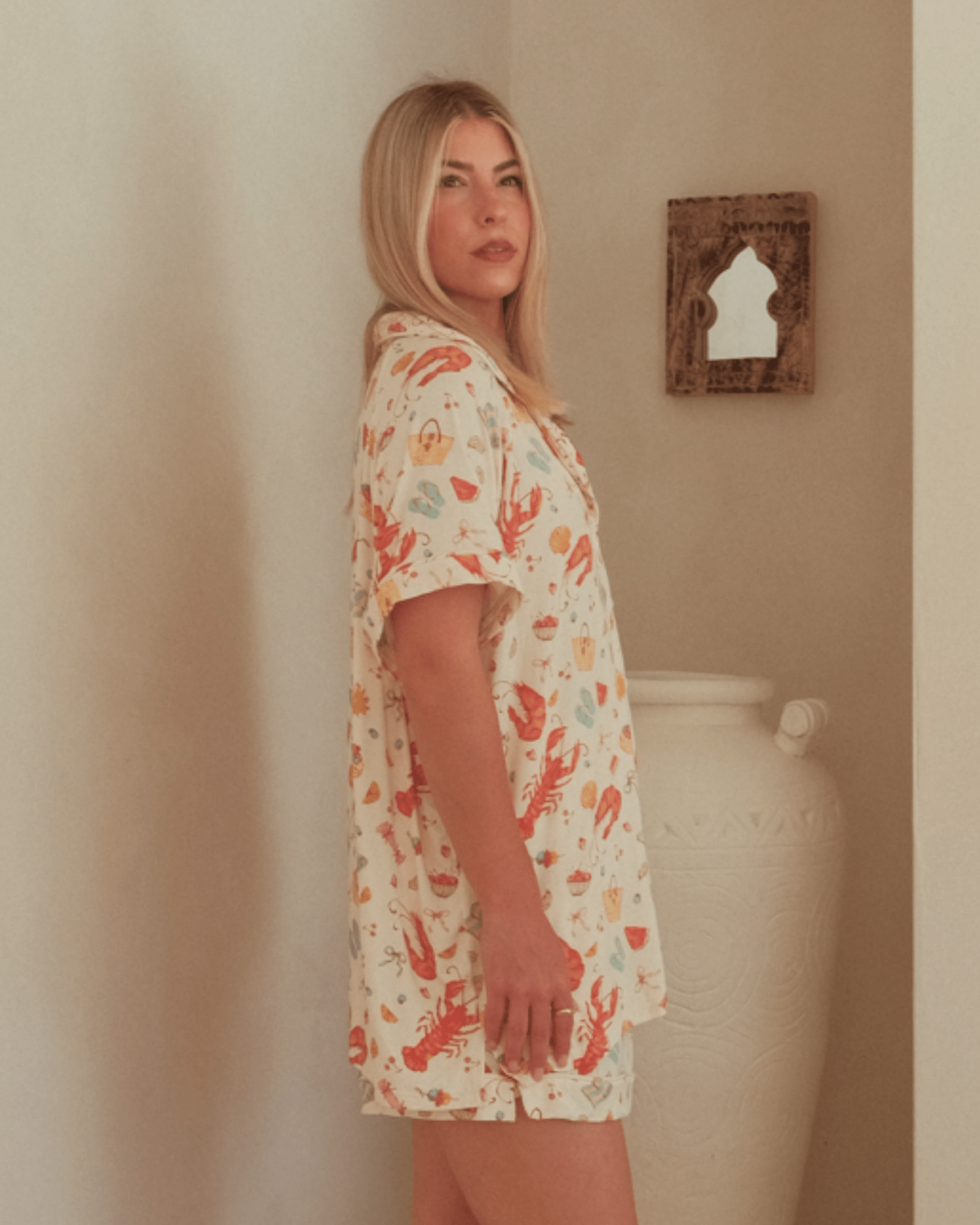 Woman wearing a pattern matching pj's  standing against a beige wall with a decorative mirror.
