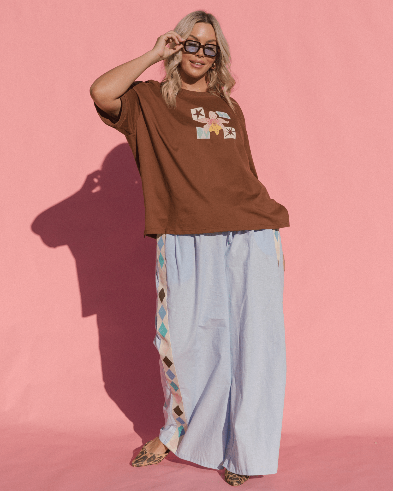 Woman wearing a brown oversized t-shirt with colorful graphics and light blue pants against a pink background