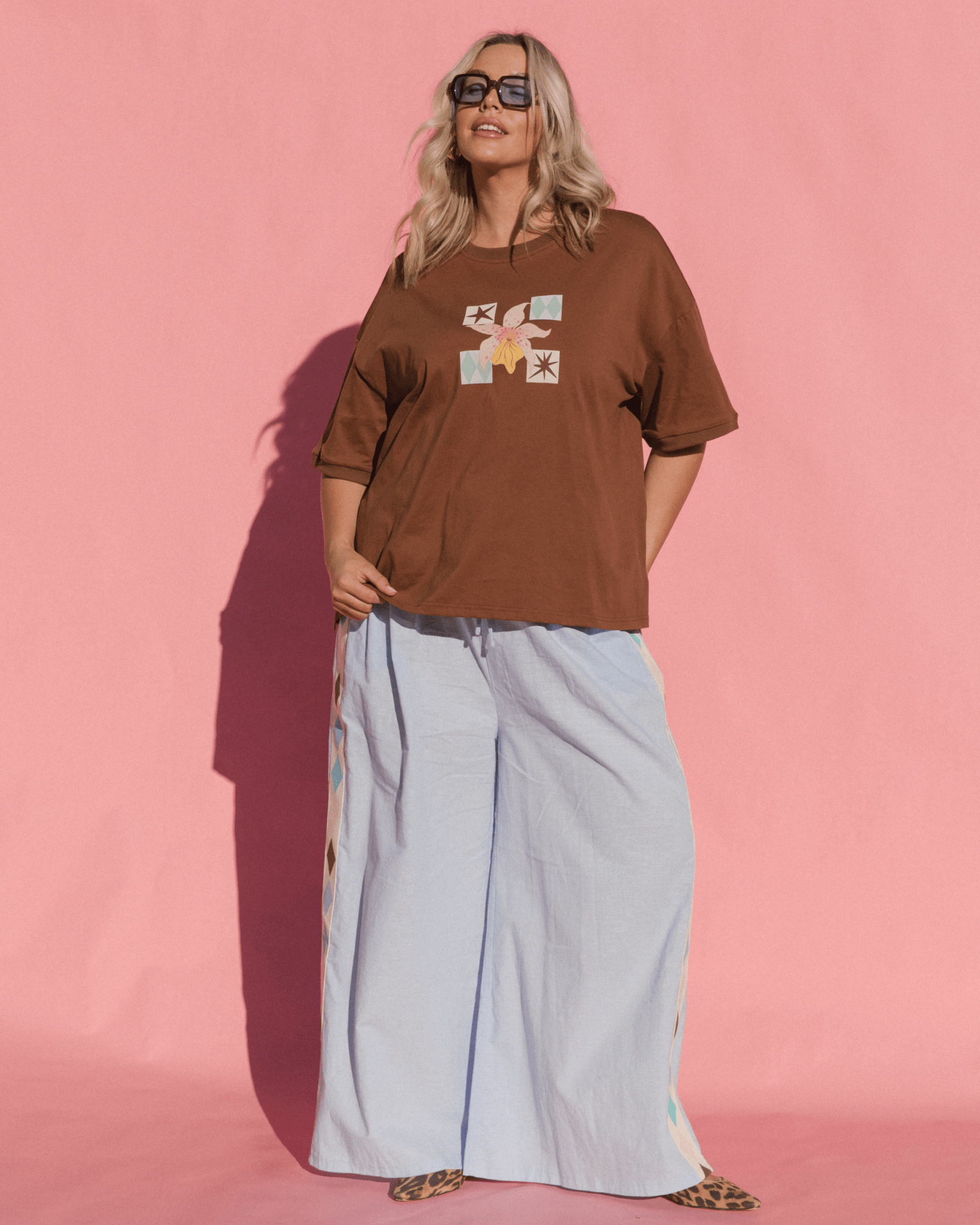 Woman wearing a brown t-shirt with graphic designs and light blue pants against a pink background
